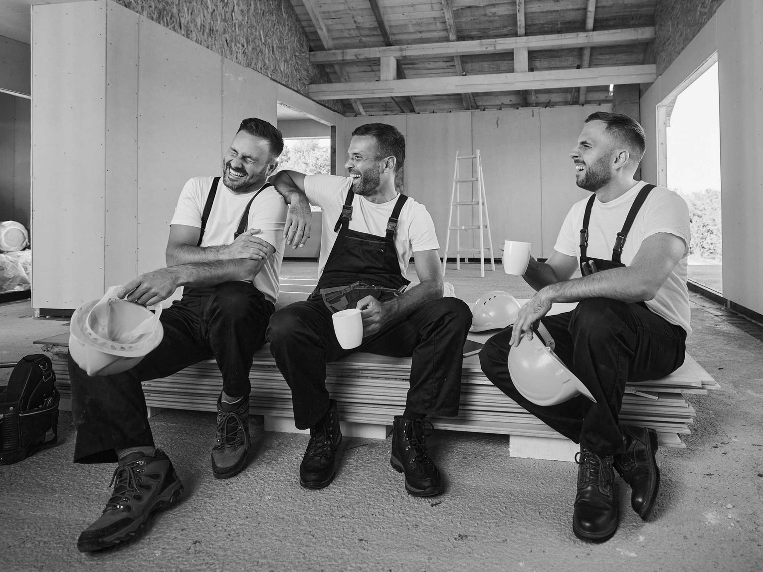 Drei Handwerker in Arbeitskleidung sitzen lachend auf einer Baustelle bei der Kaffeepause.