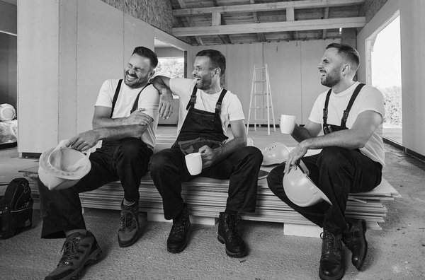 Drei Handwerker in Arbeitskleidung sitzen lachend auf einer Baustelle bei der Kaffeepause.