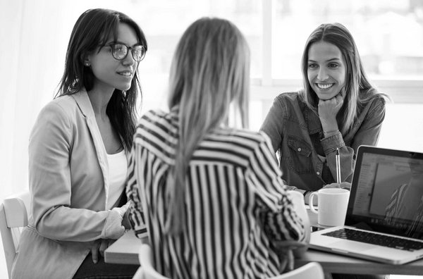 Drei Frauen in moderner Büroumgebung im Gespräch bei einem Teammeeting, Laptop und Kaffeetassen auf dem Tisch.