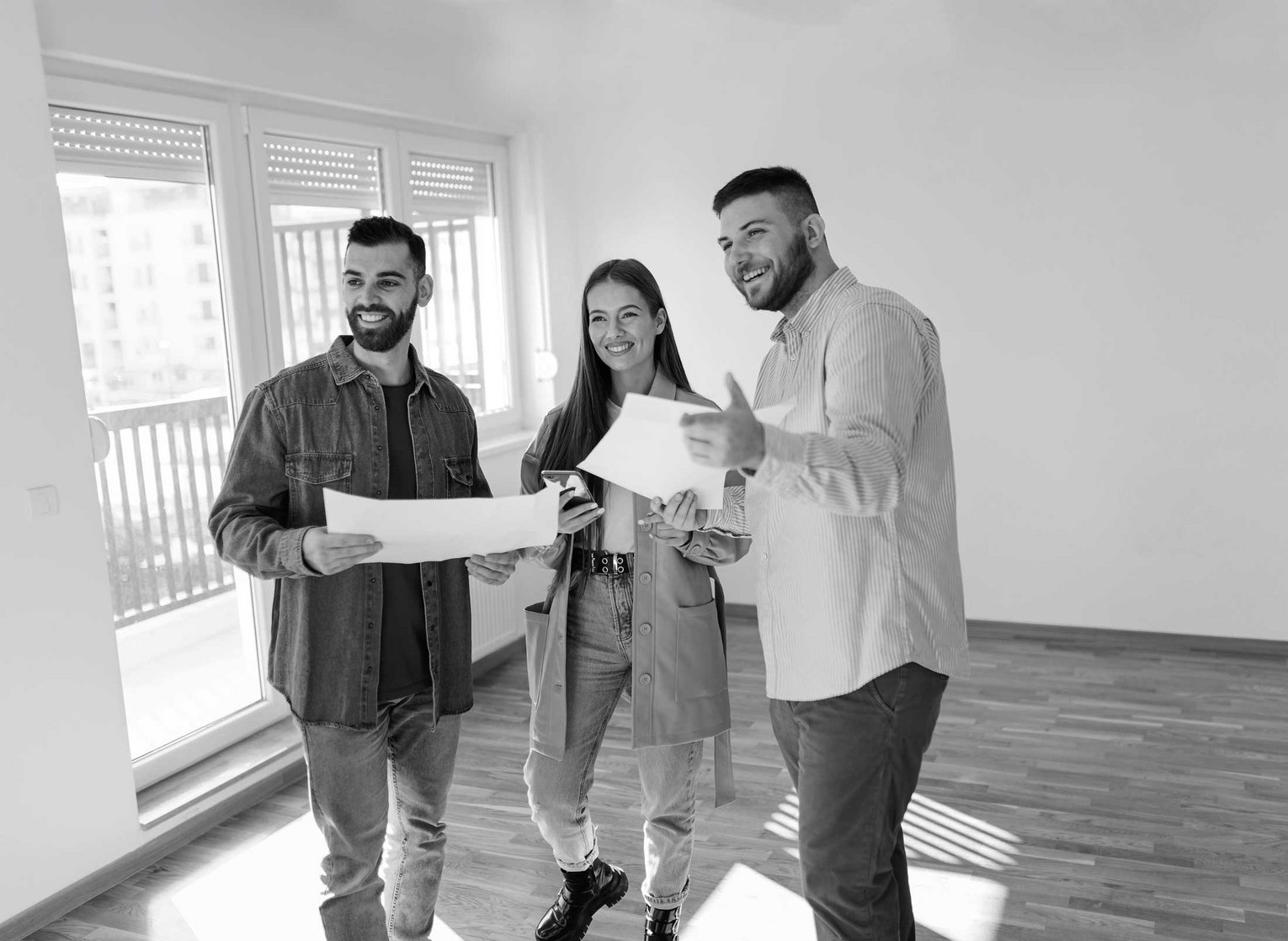 Drei junge Erwachsene in einer leeren Wohnung mit Bauplänen, vermutlich bei einer Wohnungsbesichtigung oder Planung.