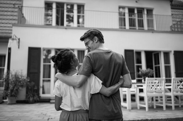Zwei Personen stehen eng umarmt im Hof vor einem Wohnhaus und schauen sich lächelnd an. Die Szene ist in Schwarz-Weiß gehalten.